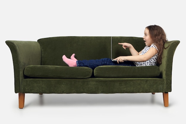 Charming fame kid in casual clothes sitting on vintage sofa indoors with notebook computer on her lap, opening mouth in astonishment and pointing finger at screen, having shocked expression