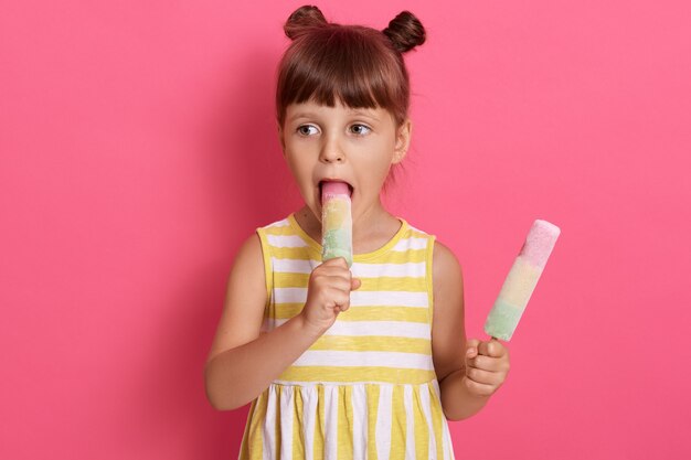 Charming female child biting water ice cream and looking aside, girl with two knots, wearing summer dress, posing isolated over pink background, stands with sorbets in hands.