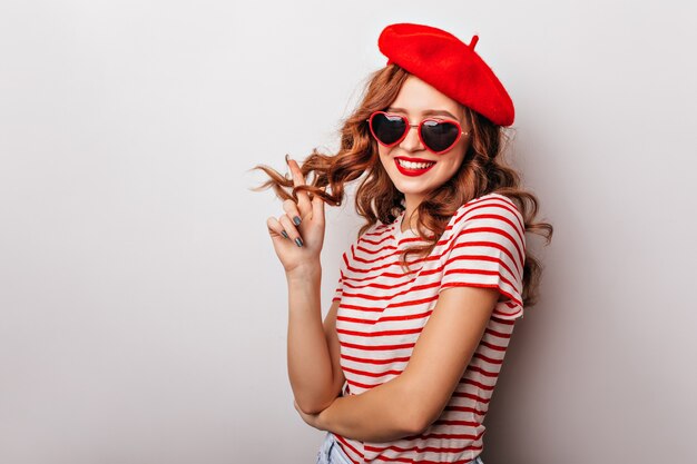 Charming female model playing with ginger curly hair. Elegant french woman in beret standing on white wall.