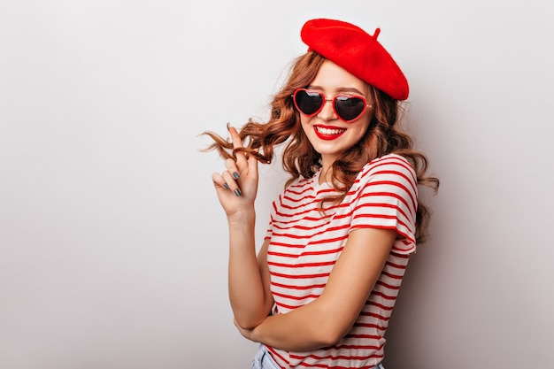 Charming female model playing with ginger curly hair. Elegant french woman in beret standing on white wall.