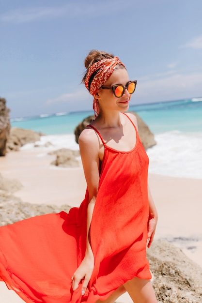 Charming female model in red dress walking down the ocean coast. Outdoor shot of enthusiastic young woman wears sunglasses during rest near sea.