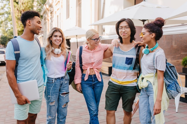 Foto gratuita affascinante studentessa con capelli biondi in bicchieri in piedi tra i compagni di classe e guardando con un sorriso al ragazzo asiatico. amici beati che discutono di esami all'aperto.