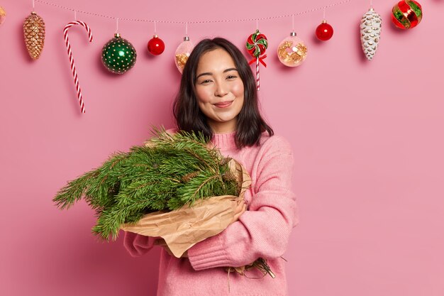 Affascinante donna con i capelli scuri e un sorriso piacevole abbraccia i rami degli abeti disposti in bouquet ha uno stato d'animo festoso indossa un maglione casual