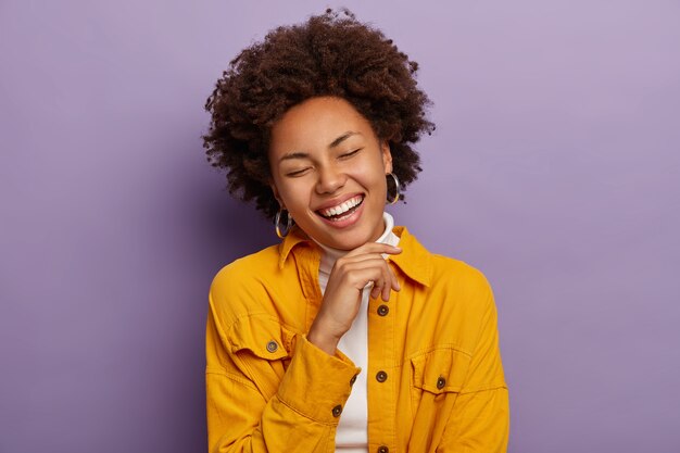 Charming feminine woman laughs from happiness, touches chin and smiles positively, feels relieved and joyful, wears fashionable yellow jacket, isolated over purple background.