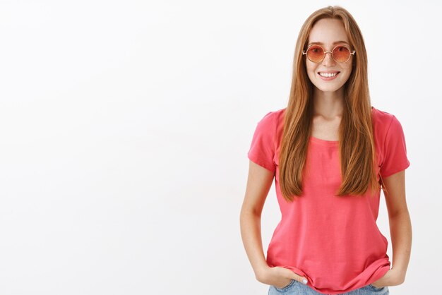 очаровательная дружелюбная девушка-хиппи с рыжими волосами и веснушками, держась за руки в карманах и небрежно улыбаясь, в модных розовых солнцезащитных очках и блузке на белой стене