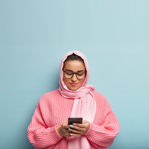 Charming friendly looking woman in hijab, follows Islam, looks attentively at tomorrow schedule, uses modern cell phone, searches information in internet, wears oversized jumper, isolated on blue wall