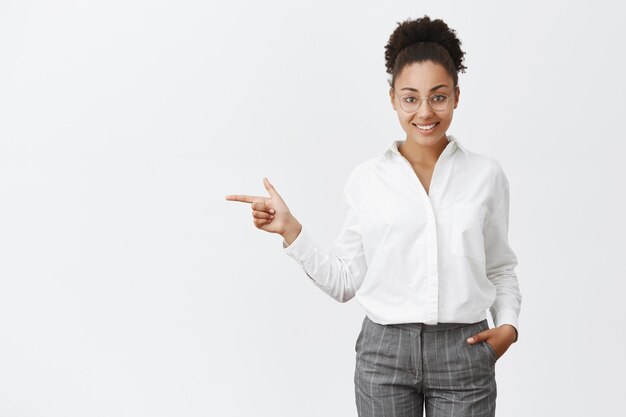 Charming and friendly office worker showing way out to customer. Portrait of polite smart and creative female employer in glasses and pants, holding hand in pocket, pointing left, indicating at exit