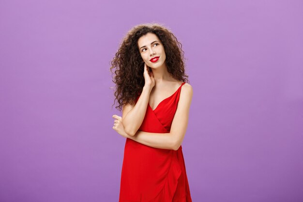 Charming and gentle elegant woman with curly hairstyle in stylish evening red dress tilting head touching neck and gazing at upper right corner tender, daydreaming posing over purple background.