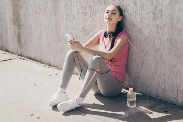 Charming girl athlete in a pink t-shirt sitting on the ground in the sun, gaining strength and energy
