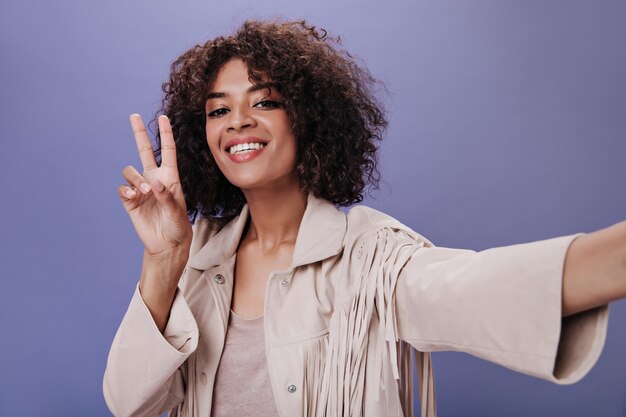 Charming girl in beige outfit smiles, takes selfie and shows sign of peace