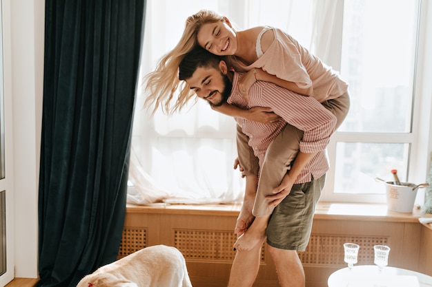 Free photo charming girl blonde jumps on back to her boyfriend. snapshot of lovers in great mood embracing.