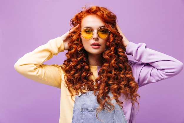 Charming girl in denim sundress and sweater touches her red hair and poses wearing glasses