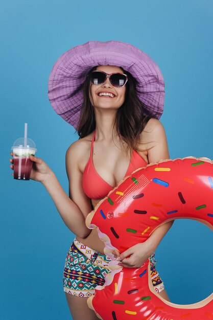 Charming girl in hat and beach dress stands with a cocktail and donut swim ring