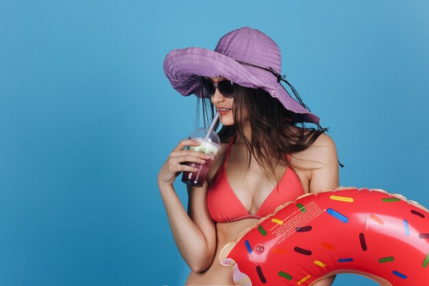 Charming girl in hat and beach dress stands with a cocktail and donut swim ring