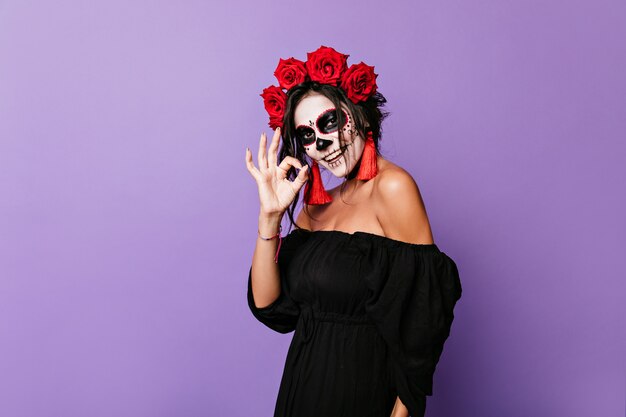 Charming girl in image of skeleton happily posing . Portrait of  cute lady in black top with red roses in curls showing OK