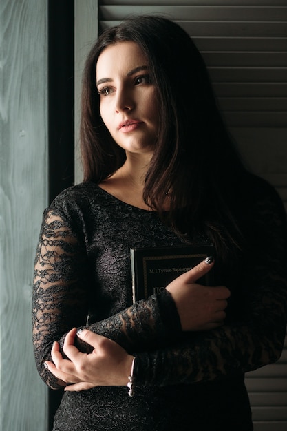 The charming girl keeps a book and stands near wall