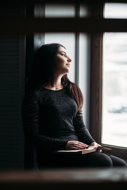 The charming girl sits on the window-sill and keeps a book