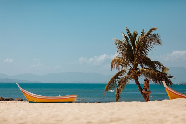 Free photo charming girl standing under palm tree leaves on sandy beach