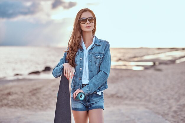 夕暮れ時の曇りのビーチでスケートボードでポーズをとるデニムのショートパンツとジャケットを着たサングラスの魅力的な女の子。