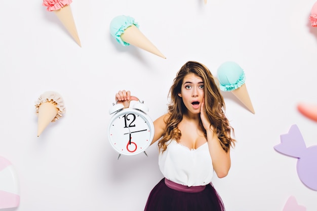 Charming girl wearing luxury dress posing with surprised face expression on cute wall with sweet decor. Portrait of worried young woman with shiny hair holding big clock.