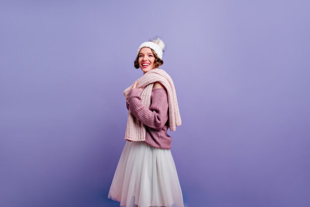 Charming girl wears cute winter hat looking with interest while posing on purple wall. Photo of good-looking cheerful lady in knitted clothes.