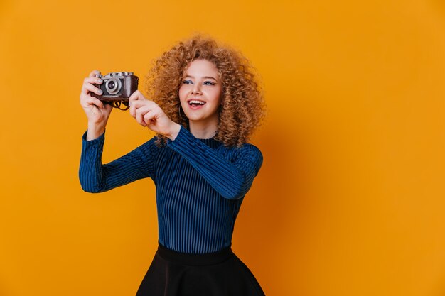 金髪のカールを持つ魅力的な女の子は、レトロなカメラで写真を作ります。黄色い空間に青いブラウスを着ている女性。