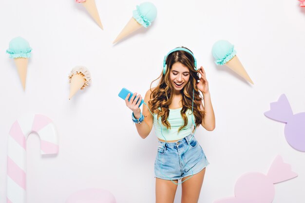 Charming girl with curly light-brown hair listening music with smile and looking down. Portrait of shapely young woman in denim shorts and earphones posing on wall decorated with ice cream.