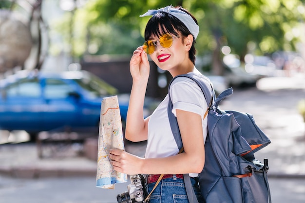 Charming girl with red lips playfully holds yellow glasses and smiling during trip around town with backpack