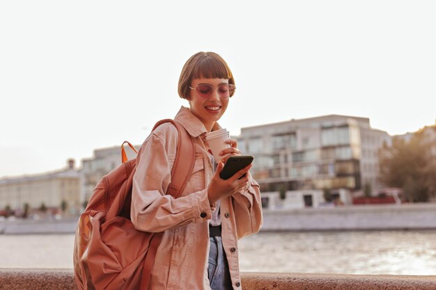 Charming girl with short hair holding phone on backdrop of river Woman in glasses and denim jacket with backpack smiles and holds cup of tea in city