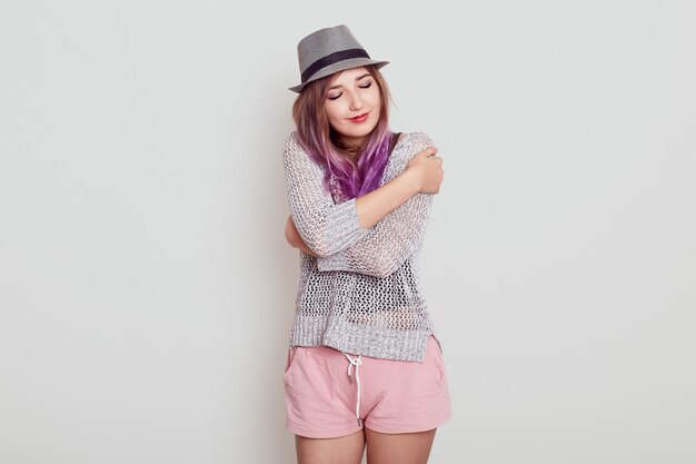 Charming gorgeous European woman keeps eyes closed, smiles with pleasure, feels comfort, hugs herself, wearing hat, shirt and short, isolated over grey background.