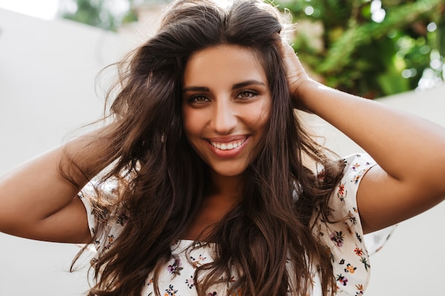 Charming green-eyed woman touches her long dark hair and smiles looking at front on white wall with plants