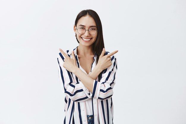 Charming happy female in glasses and striped blouse, crossing hands over chest and pointing left and right while smiling joyfully, posing over gray wall