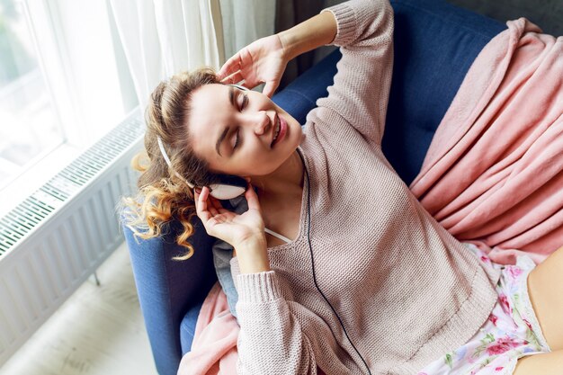 Charming happy girl with  blonde wavy hairstyle lying on blue sofa  on her living room and looking up. Cute  girl showing signs end  enjoying favorite music.