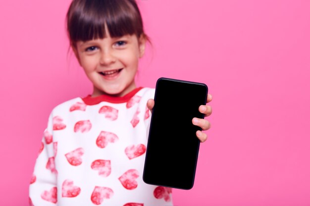 Charming happy smiling little girl wearing casual style jumper and showing blank screen of mobile phone in her hand, posing isolated over pink wall.