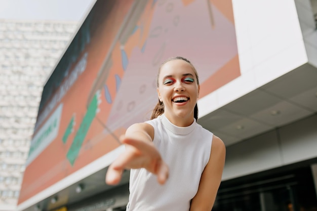 Charming happy woman with bright make up is reaching for the camera and laughing on the city background