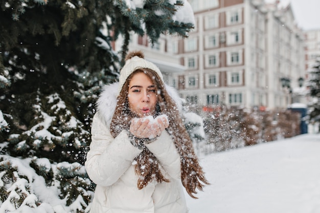 Charming joyful woman blowing snowflakes from her hands in winter day outdoor on street.