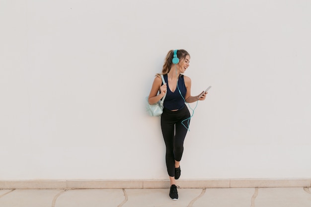 Charming joyful young woman in sportswear enjoying chatting on phone, lovely music in headphones.  Outwork, sportive lifestyle, fashionable model, time for fitness, having fun