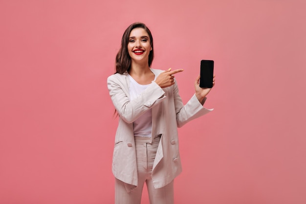 ベージュのスーツを着た魅力的な女性は、ピンクの背景にスマートフォンを示しています。白い服を着た明るいメイクのスタイリッシュな女性が彼女の携帯電話に表示されます。