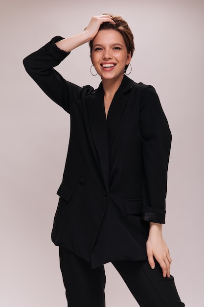 Charming lady in black classic outfit smiling on isolated background. Attractive woman in dark suit laughing and touching hair