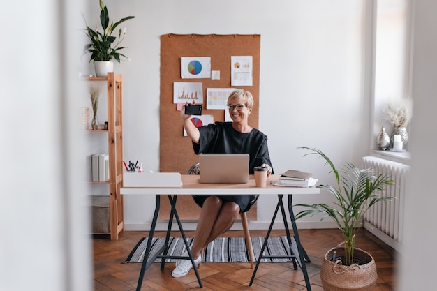 Charming lady in black dress takes selfie in office