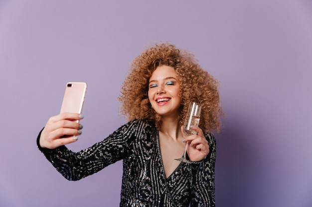 Charming lady in black shiny top laughs, closing her eyes, holding glass of champagne and making selfie on purple space.