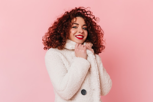 Charming lady dressed in white coat looking at camera. portrait of dark-haired girl with blue eyes posing on pink space.