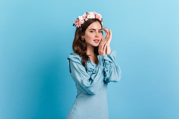 Charming lady in elegant blue dress posing on isolated wall. Indoors portrait of young woman with pink flowers in dark hair.