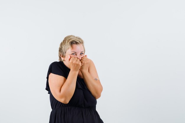 Charming lady holding fists on her mouth in black blouse and looking cheery. front view.