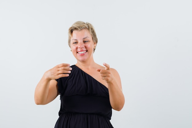 Charming lady holding something while pointing at camera in black blouse and looking pleased 