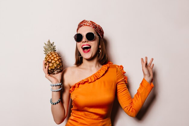 Charming lady in orange blouse, headdress and glasses laughs and holds pineapple on white space.