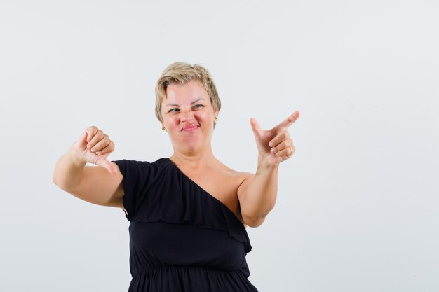 Charming lady posing like holding phone while showing thumb down in black blouse and looking displeased. front view.