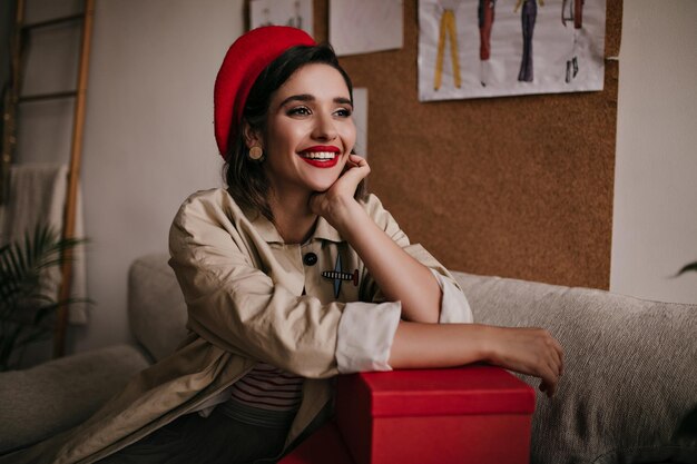 赤いスタイリッシュなベレー帽の魅力的な女性が笑顔でソファに座っているベージュのコートのポーズで黒髪の素晴らしい若い女性
