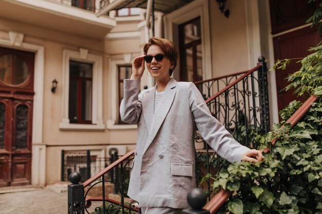 Charming lady in stylish suit takes off her sunglasses and walks outside. Young woman in grey jacket and pants smiling opposite building