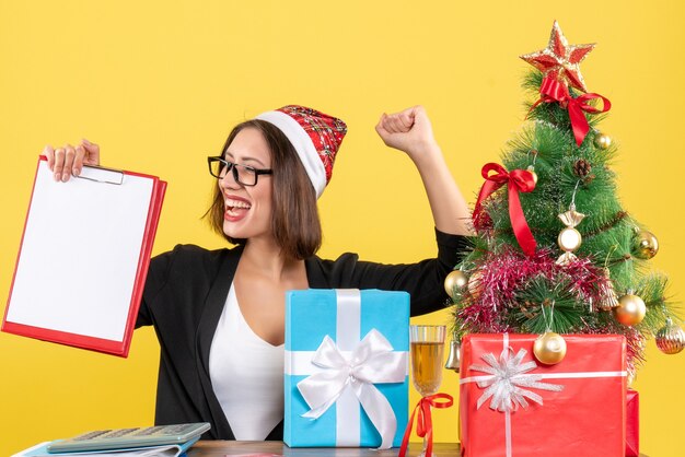 Charming lady in suit with santa claus hat and eyeglasses holding documents proudly in the office 